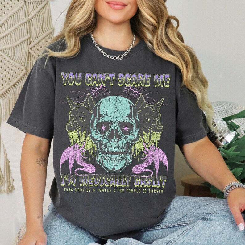 May include: Dark gray t-shirt with a graphic design featuring a teal skull, two wolf-like creatures, and text that reads "YOU CAN'T SCARE ME I'M MEDICALLY GASLIT. THIS BODY IS A TEMPLE & THE TEMPLE IS CURSED."