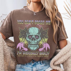 May include: Brown t-shirt with a graphic design featuring a skull, two wolf-like creatures, and text that reads "YOU CAN'T SCARE ME I'M MEDICALLY GASLIT THIS BODY IS A TEMPLE & THE TEMPLE IS CURSED."