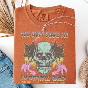 May include: Burnt orange t-shirt with a graphic design featuring a skull, two snarling wolf-like creatures, and two winged serpent-like creatures. The text "YOU CAN'T SCARE ME I'M MEDICALLY GASLIT" is printed on the shirt. A silver necklace is draped across the shirt.