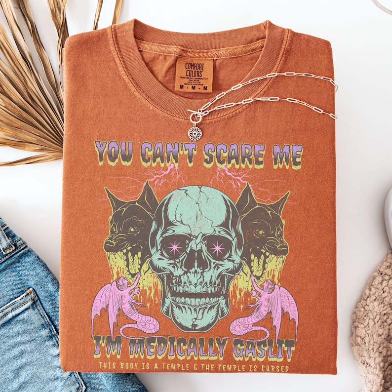May include: Burnt orange t-shirt with a graphic design featuring a skull, two snarling wolf-like creatures, and two winged serpent-like creatures. The text "YOU CAN'T SCARE ME I'M MEDICALLY GASLIT" is printed on the shirt. A silver necklace is draped across the shirt.