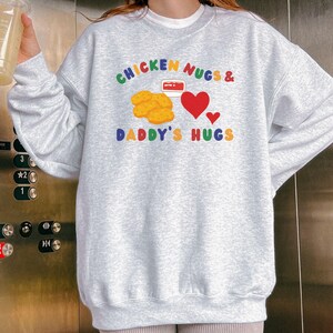 May include: A light gray sweatshirt with the text "CHICKEN NUGS & DADDY'S HUGS" in colorful letters. The design includes illustrations of chicken nuggets, a container of dipping sauce, and red hearts. The sweatshirt has a crew neck.
