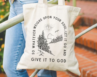 May include: A white canvas tote bag with a black graphic design. The graphic features a cross, a bird, and trees. The text on the bag reads "So whatever weighs upon your soul, let it go and give it to God."