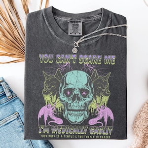May include: Dark gray Comfort Colors t-shirt with a graphic skull design. The design includes the text "YOU CAN'T SCARE ME I'M MEDICALLY GASLIT" and other phrases. A silver necklace with a pendant is draped over the shirt.