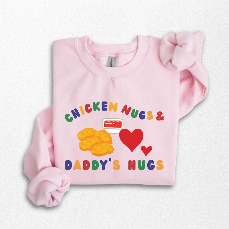 May include: A light pink sweatshirt with the text "CHICKEN NUGS & DADDY'S HUGS" in colorful letters. The design includes illustrations of chicken nuggets, a dipping sauce, and red hearts.
