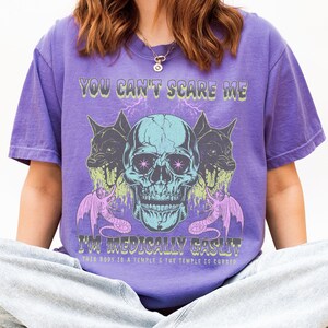 May include: Purple t-shirt with a graphic design featuring a teal skull, two snarling black dogs, and text that reads "YOU CAN'T SCARE ME I'M MEDICALLY GASLIT THIS BODY IS A TEMPLE & THE TEMPLE IS CURSED."