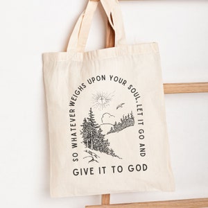 May include: A white canvas tote bag with black text and a graphic of a forest scene. The text reads "SO WHATEVER WEIGHS UPON YOUR SOUL, LET IT GO AND GIVE IT TO GOD".