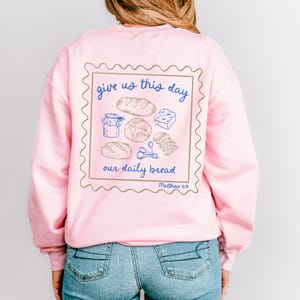 May include: Pink sweatshirt with a graphic of a stamp with the text "give us this day our daily bread Matthew 6:11" and a drawing of bread, a jar, and other food items.
