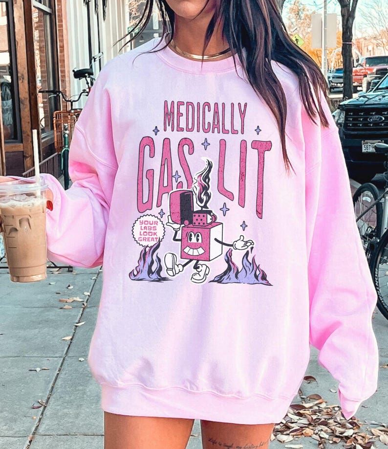 Pode incluir: Camisola cor-de-rosa com um gr&aacute;fico de um isqueiro de desenho animado com o texto "Medically Gaslit" e "Your Labs Look Great!" impresso nela.