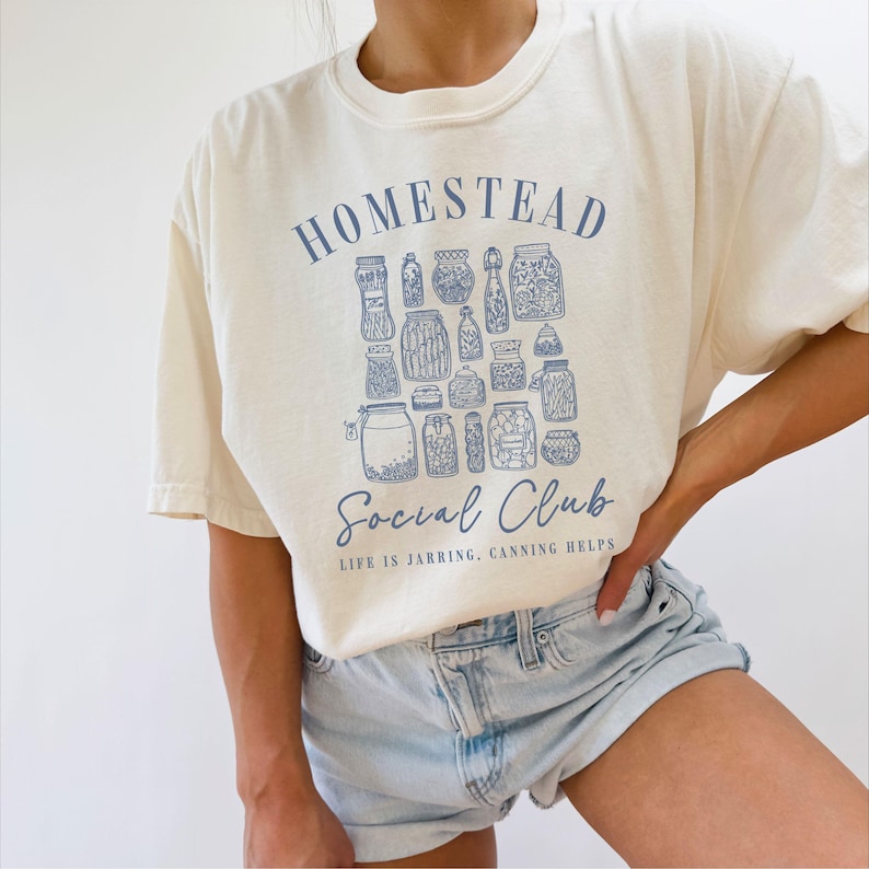 May include: A cream-colored t-shirt with a blue graphic of jars and the text "Homestead Social Club. Life is Jarring. Canning Helps."