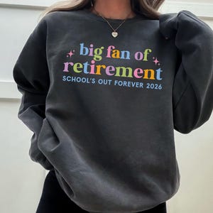 May include: Charcoal grey sweatshirt featuring the phrase "big fan of retirement" in vibrant colours. The text "SCHOOL'S OUT FOREVER 2026" is printed below. The sweatshirt has a crew neck and long sleeves.