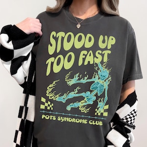 May include: Dark gray t-shirt with the text "STOOD UP TOO FAST" in light green. A turquoise skeleton graphic with flames is featured below the text. The words "POTS SYNDROME CLUB" are printed at the bottom.