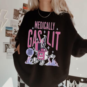Pode incluir: Camisola de moletom preta com um gr&aacute;fico rosa de um isqueiro de desenho animado com o texto "Medically Gaslit" e um c&iacute;rculo menor com o texto "Your Labs Look Great".