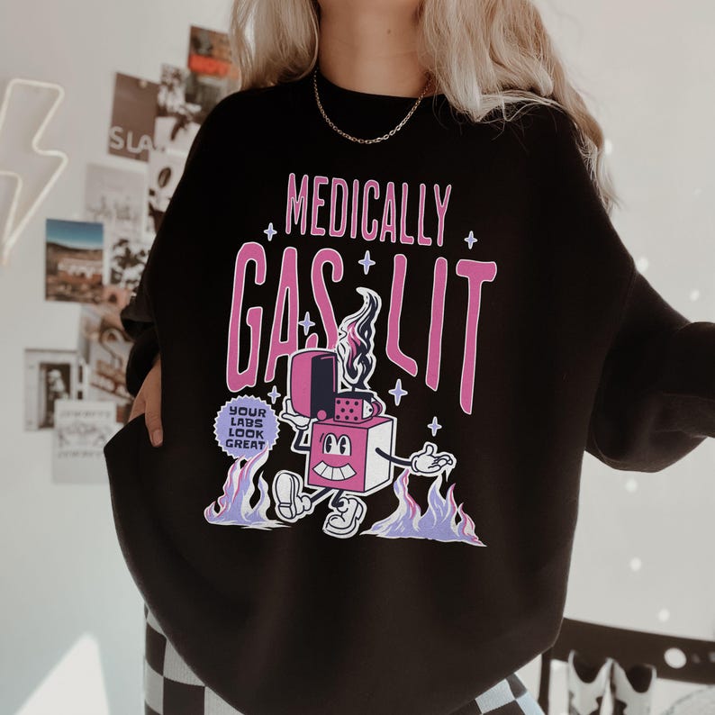 Pode incluir: Camisola de moletom preta com um gr&aacute;fico rosa de um isqueiro de desenho animado com o texto "Medically Gaslit" e um c&iacute;rculo menor com o texto "Your Labs Look Great".