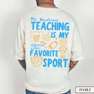 May include: Ivory-colored t-shirt with a blue and orange graphic. The graphic includes the text "Mr. Henderson TEACHING IS MY FAVORITE SPORT" along with illustrations of sports equipment like a basketball hoop, soccer ball, and baseball.