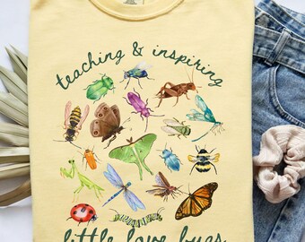 May include: A light yellow t-shirt with a watercolor illustration of various insects, including butterflies, bees, and dragonflies. The text "teaching & inspiring" and "little love bugs" are printed in green script.