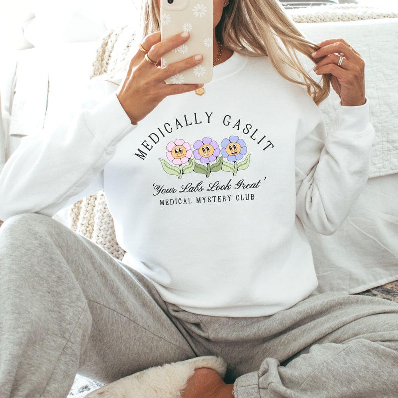 May include: White sweatshirt with the text "Medically Gaslit" and a graphic of three cartoon flowers with faces. The text below the flowers reads "Your Labs Look Great" and "Medical Mystery Club".