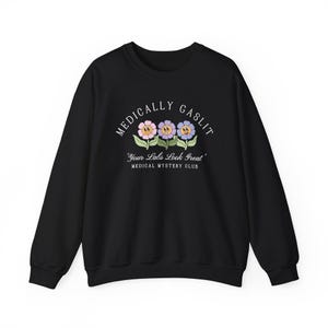 May include: Black sweatshirt with a white graphic print of three cartoon flowers with smiling faces. The text reads "Medically Gaslit" and "Your Labs Look Great" with the words "Medical Mystery Club" below.