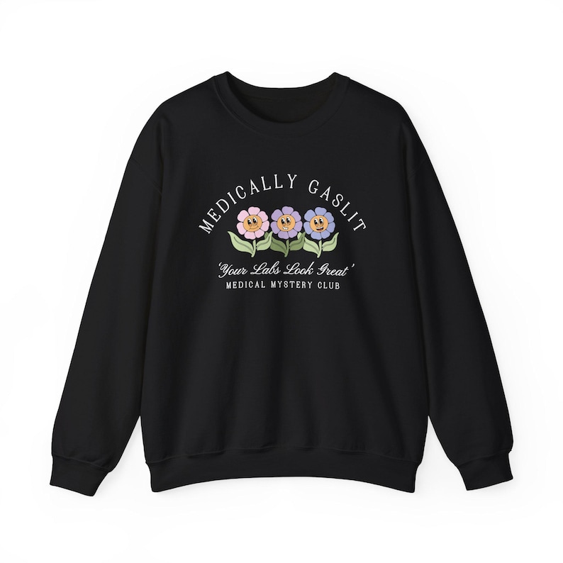 May include: Black sweatshirt with a white graphic print of three cartoon flowers with smiling faces. The text reads "Medically Gaslit" and "Your Labs Look Great" with the words "Medical Mystery Club" below.