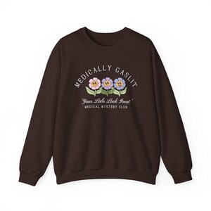 May include: A brown sweatshirt with the text "Medically Gaslit" in white, featuring three cartoon flowers with smiling faces. The text below the flowers reads "Your Labs Look Great" and "Medical Mystery Club".