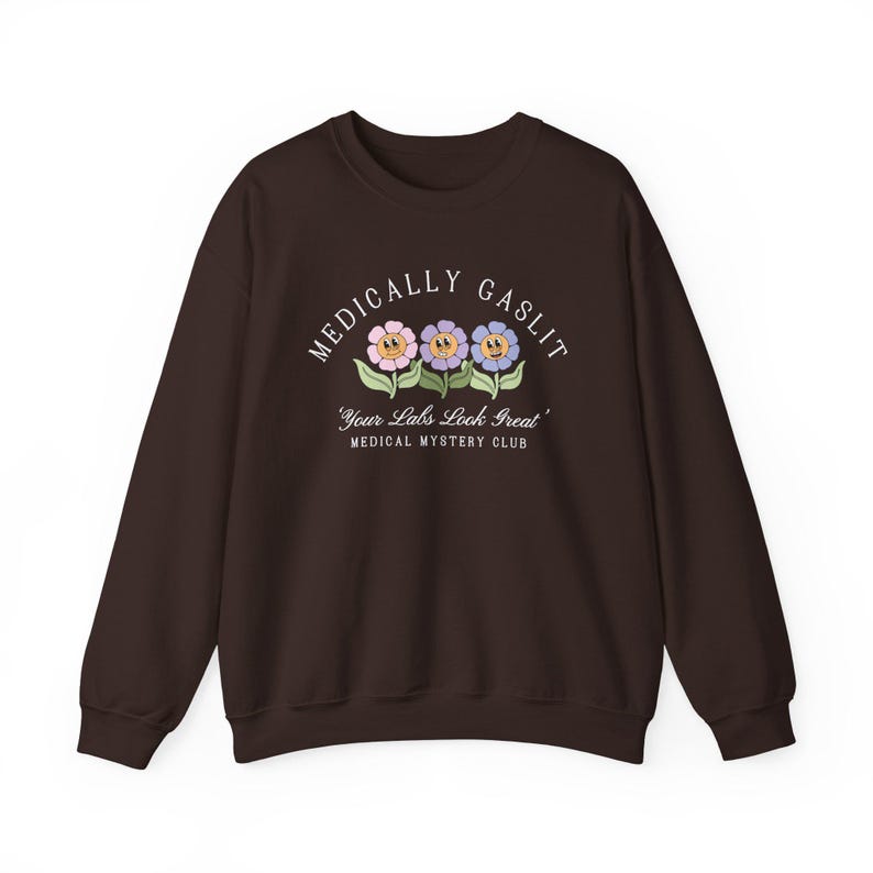 May include: A brown sweatshirt with the text "Medically Gaslit" in white, featuring three cartoon flowers with smiling faces. The text below the flowers reads "Your Labs Look Great" and "Medical Mystery Club".