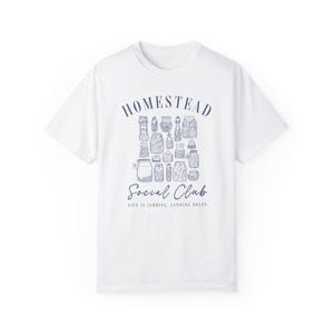 May include: White t-shirt with a blue graphic design that says "Homestead Social Club" and "Life is Jarring, Canning Helps". The graphic features a collection of jars with different contents.