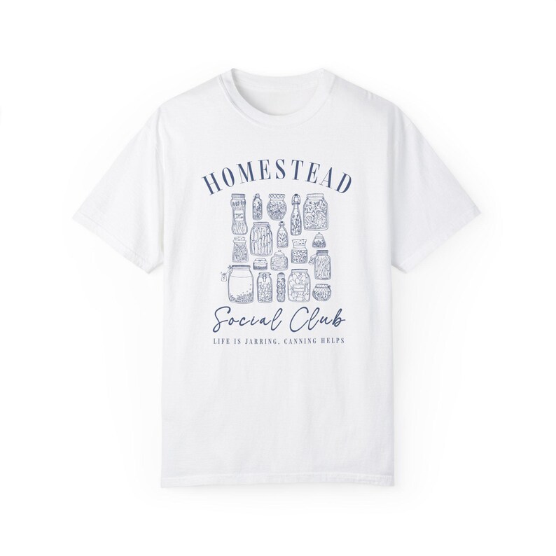 May include: White t-shirt with a blue graphic design that says "Homestead Social Club" and "Life is Jarring, Canning Helps". The graphic features a collection of jars with different contents.