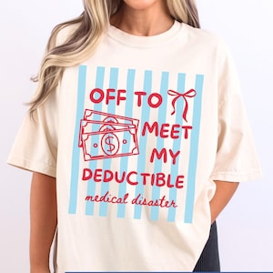 May include: Cream-colored t-shirt with a graphic design. The design features red text and illustrations on a light blue striped background. The text reads "OFF TO MEET MY DEDUCTIBLE medical disaster."