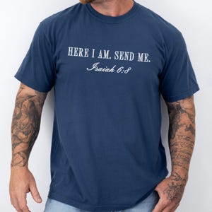 May include: A navy blue t-shirt featuring the white text "HERE I AM. SEND ME." and "Isaiah 6:8". The t-shirt has a crew neck and is likely made from a soft fabric, suitable for casual wear.