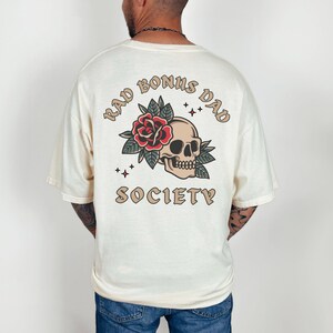 May include: A cream colored t-shirt with a graphic print of a skull with a rose and leaves. The text "Rad Bonus Dad Society" is printed in a brown font around the skull.
