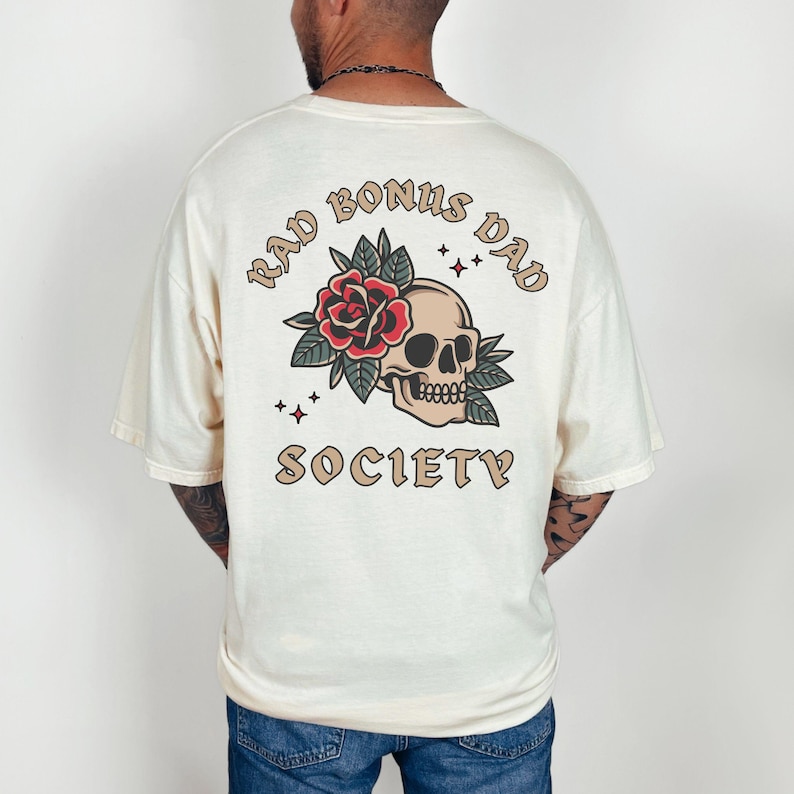 May include: A cream colored t-shirt with a graphic print of a skull with a rose and leaves. The text "Rad Bonus Dad Society" is printed in a brown font around the skull.