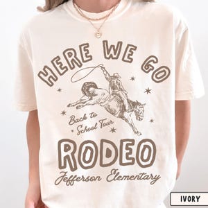 May include: An ivory short-sleeve t-shirt featuring brown text that reads HERE WE GO, Back to School Tour, RODEO, and Jefferson Elementary. The graphic includes a line-art illustration of a cowboy riding a bucking horse with a lasso.