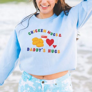 May include: Light blue sweatshirt with the text "CHICKEN NUGS & DADDY'S HUGS" in rainbow colors. The graphic includes illustrations of chicken nuggets, a container of dipping sauce, and red hearts. A casual, comfortable top.