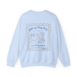 May include: Light blue sweatshirt with a graphic of a stamp with the text "give us this day our daily bread" and a drawing of bread and other food items. The text "Matthew 6:11" is below the stamp.