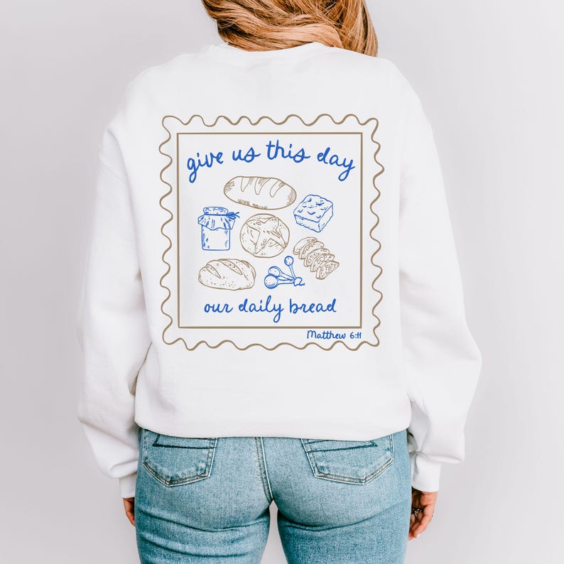 May include: White sweatshirt with a brown and blue graphic of a stamp with the text "give us this day our daily bread Matthew 6:11" and a drawing of bread and other food items.