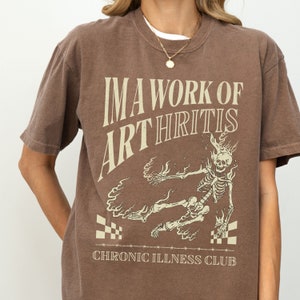 Pu&ograve; includere: Maglietta marrone con un grafico di uno scheletro in fiamme e il testo "I'm a work of arthritis Chronic Illness Club".