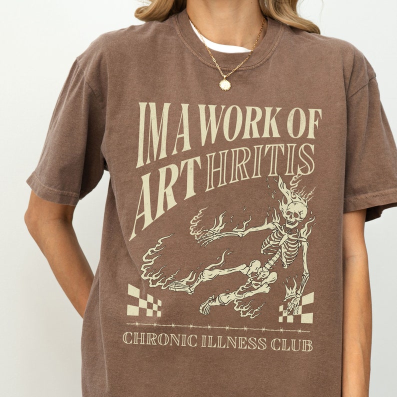 Pu&ograve; includere: Maglietta marrone con un grafico di uno scheletro in fiamme e il testo "I'm a work of arthritis Chronic Illness Club".