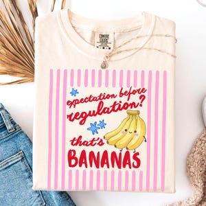May include: Cream-colored t-shirt with the phrase "expectation before regulation? that's BANANAS" in red and pink. The design includes a bunch of smiling bananas and blue star accents. The shirt is a Comfort Colors brand.