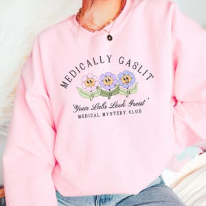 May include: Pink sweatshirt with the text "Medically Gaslit" and a graphic of three cartoon flowers with faces. The text below the flowers reads "Your Labs Look Great, Medical Mystery Club."