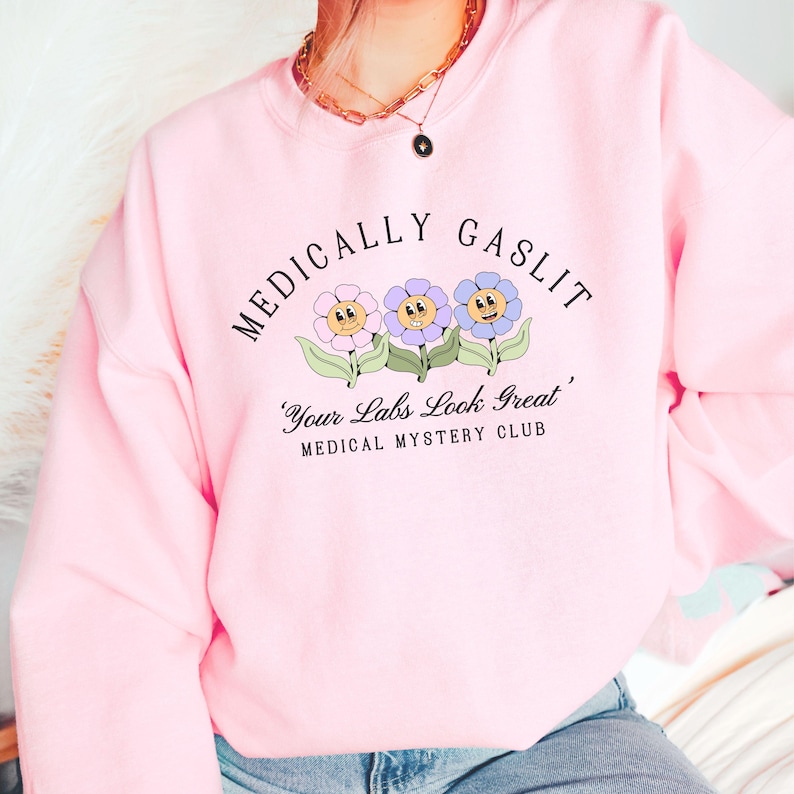 May include: Pink sweatshirt with the text "Medically Gaslit" and a graphic of three cartoon flowers with faces. The text below the flowers reads "Your Labs Look Great, Medical Mystery Club."