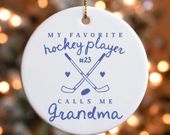 May include: White ceramic round ornament with blue text and graphics. The text reads "MY FAVORITE hockey player #23 CALLS ME Grandma." Features crossed hockey sticks, a puck, and hearts.