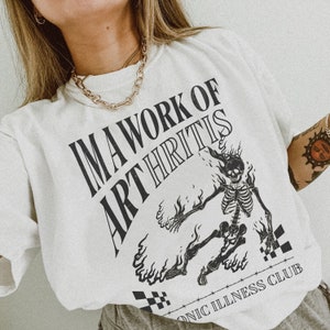 Pu&ograve; includere: Maglietta bianca con una grafica nera di uno scheletro in fiamme con il testo "I'm a work of arthritis" e "Chronic Illness Club".