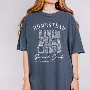 May include: A navy blue t-shirt with a white graphic of jars and the text "Homestead Social Club. Life is Jarring, Canning Helps."