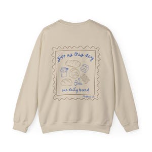 May include: A beige sweatshirt with a graphic print of a stamp with the text "give us this day our daily bread" and a drawing of bread and other food items. The text "Matthew 6:11" is printed below the stamp.