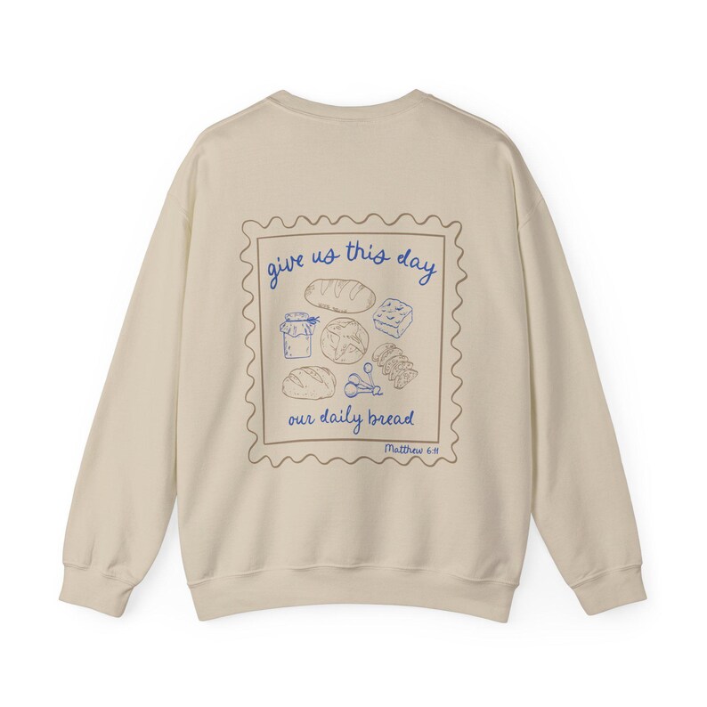 May include: A beige sweatshirt with a graphic print of a stamp with the text "give us this day our daily bread" and a drawing of bread and other food items. The text "Matthew 6:11" is printed below the stamp.