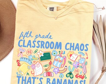以下が含まれることがあります： バターイエローのTシャツで、青い文字で「fifth grade CLASSROOM CHAOS THAT'S BANANAS!」と書かれ、バックパック、ハサミ、黒板、鉛筆、A+の評価がついた紙などの学用品のカラフルなグラフィックプリントが施されています。