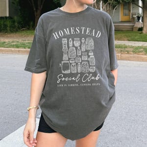 May include: A dark gray t-shirt with a white graphic design featuring a collection of jars and the text "Homestead Social Club. Life is Jarring, Canning Helps."