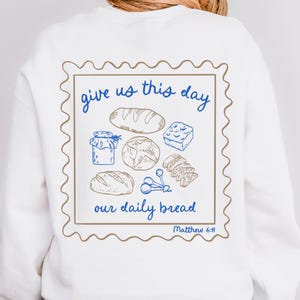 May include: White sweatshirt with a brown and blue graphic of a stamp with the text "give us this day our daily bread Matthew 6:11" and illustrations of bread, a jar, and other food items.