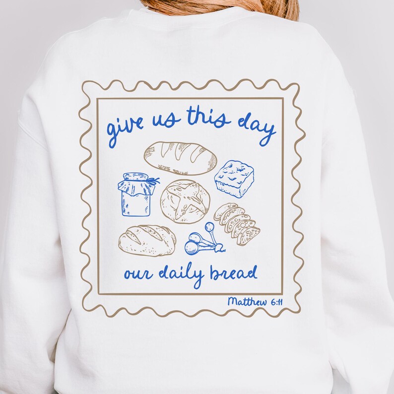 May include: White sweatshirt with a brown and blue graphic of a stamp with the text "give us this day our daily bread Matthew 6:11" and illustrations of bread, a jar, and other food items.