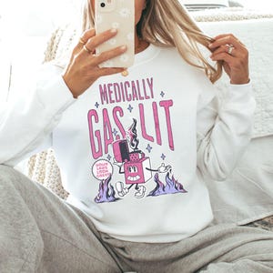 Pode incluir: Camisola branca com um gr&aacute;fico rosa de um isqueiro de desenho animado com o texto "Medically Gaslit" e "Your Labs Look Great!"