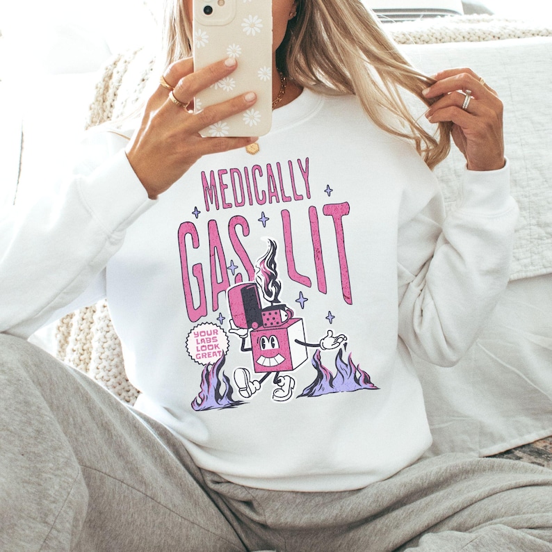 Pode incluir: Camisola branca com um gr&aacute;fico rosa de um isqueiro de desenho animado com o texto "Medically Gaslit" e "Your Labs Look Great!"